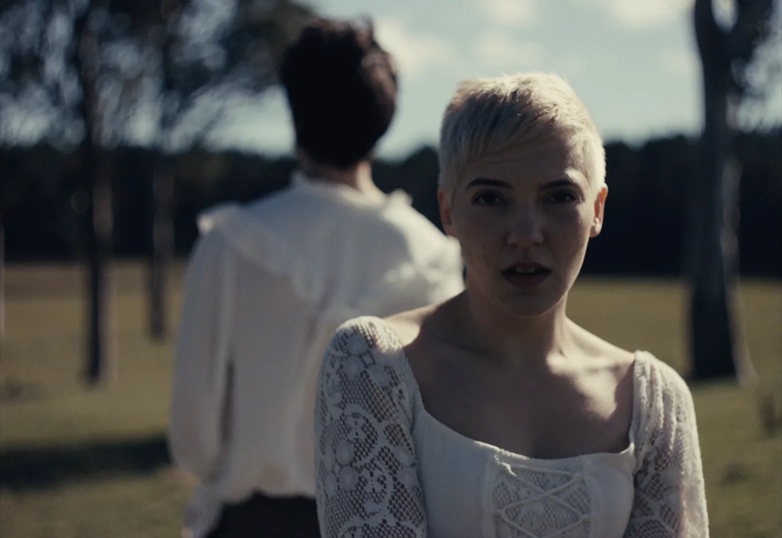 A woman with short bleached hair and a lace blouse looks straight down the lens in a sunlit paddock, a second figure soft behind her.