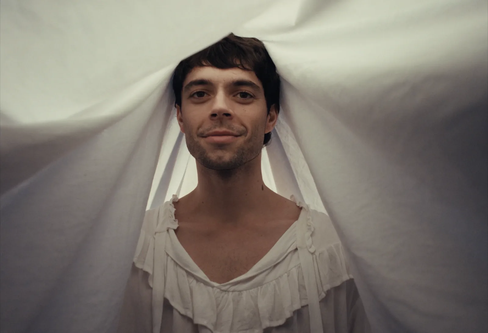 A smiling man in a ruffled white shirt pushes his head up through a white sheet, lit soft and warm.